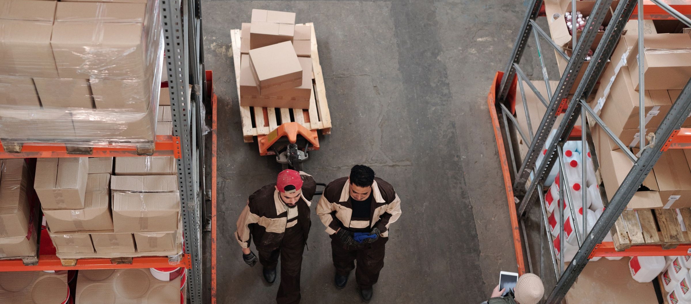 Due operatori che preparano spedizioni fra le scaffalature, con cartoni e pallet fasciati.