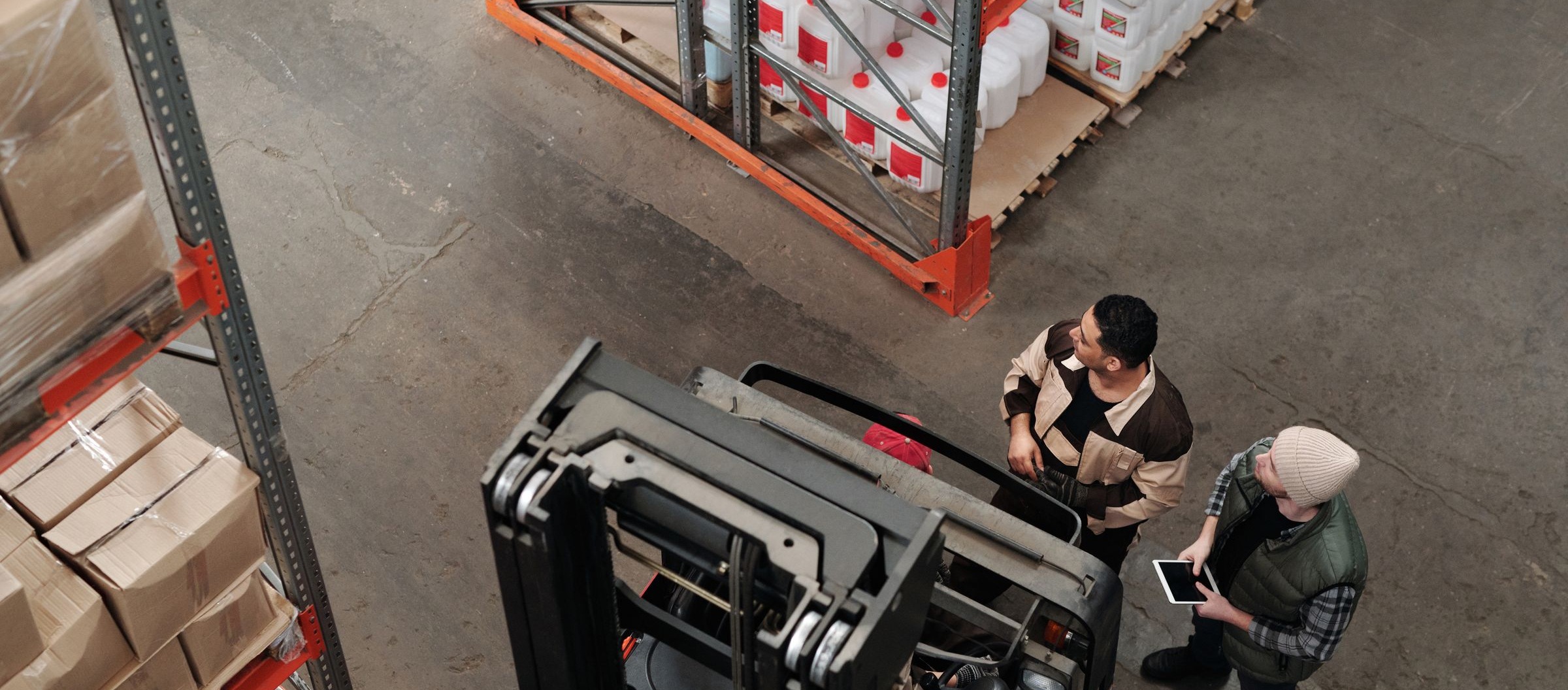 Vista dall'alto di due operatori accanto a un carrello elevatore, fra scaffalature e merce su pallet.