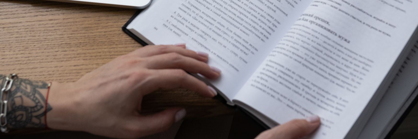 Una mano tiene aperte le pagine di un libro su una scrivania di legno, accanto a un portatile.