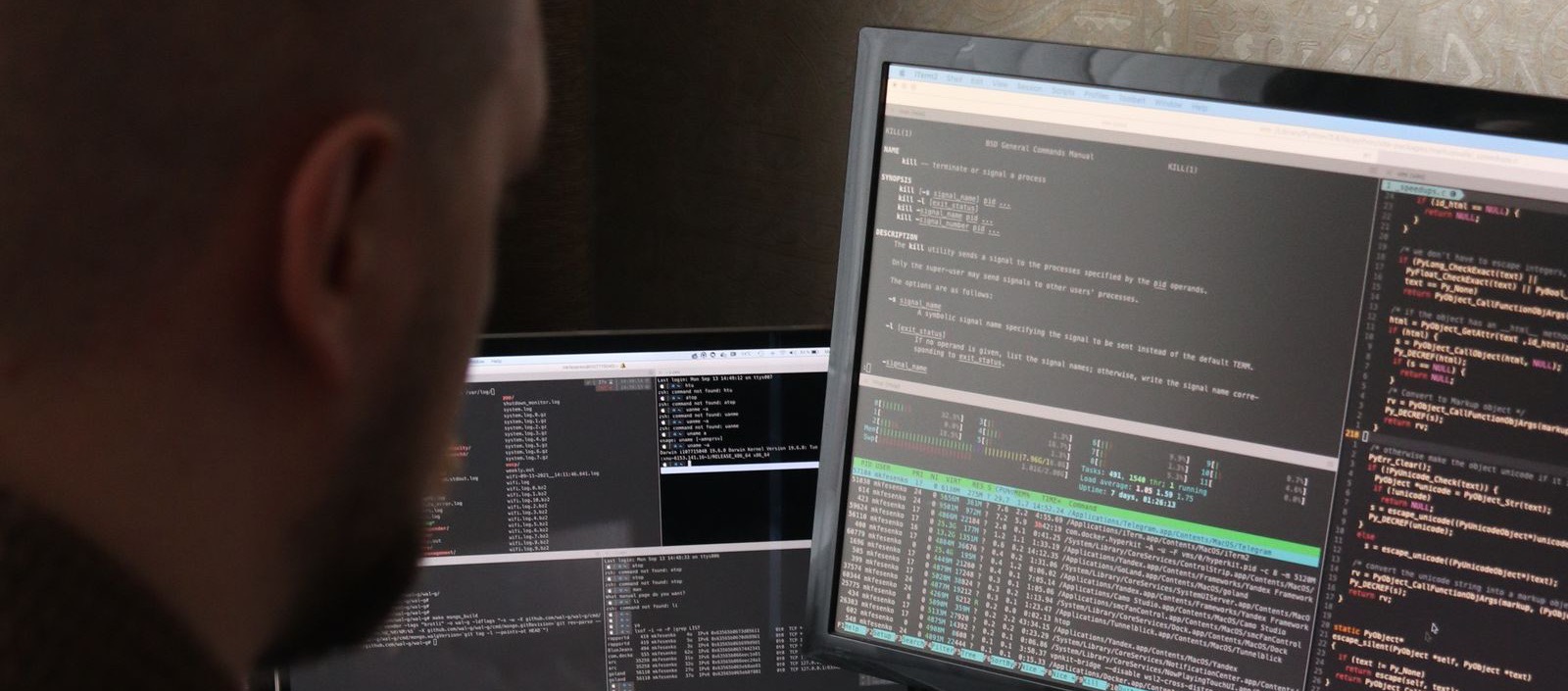 Una persona di spalle davanti a tre schermi con un manuale di sistema, un monitor di processi e del codice.